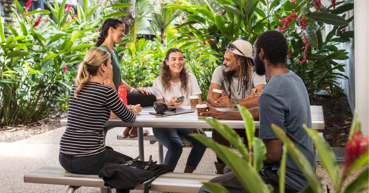 A group of five people enjoy a friendly conversation over coffee at an outdoor table surrounded by lush greenery