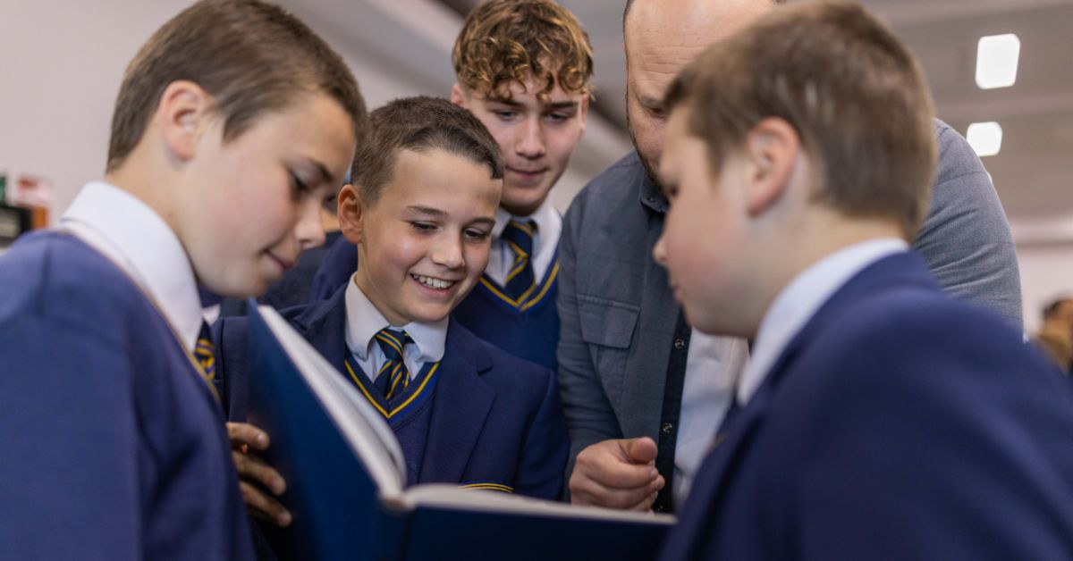 A group of young boys in blue school blazers, gathered closely around an open book, showing expressions of curiosity and joy. A male teacher, partially bald and dressed in a gray jacket, leans in attentively from behind. The boys and teacher are engaged in a discussion, focused on the content of the book, in a brightly lit classroom setting